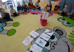 Dziewczynka podnosi ilustrację z grzebieniem, w tle siedzą na dywanie dzieci.